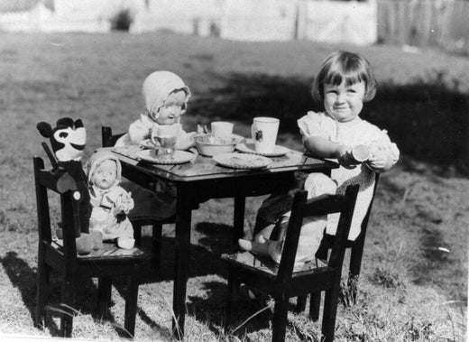 Black and white vintage photograph of a young girl having a tea party outdoors with two dolls, including a Topsy-Turvy reversible doll on a chair, and a toy tea set arranged on a small table.